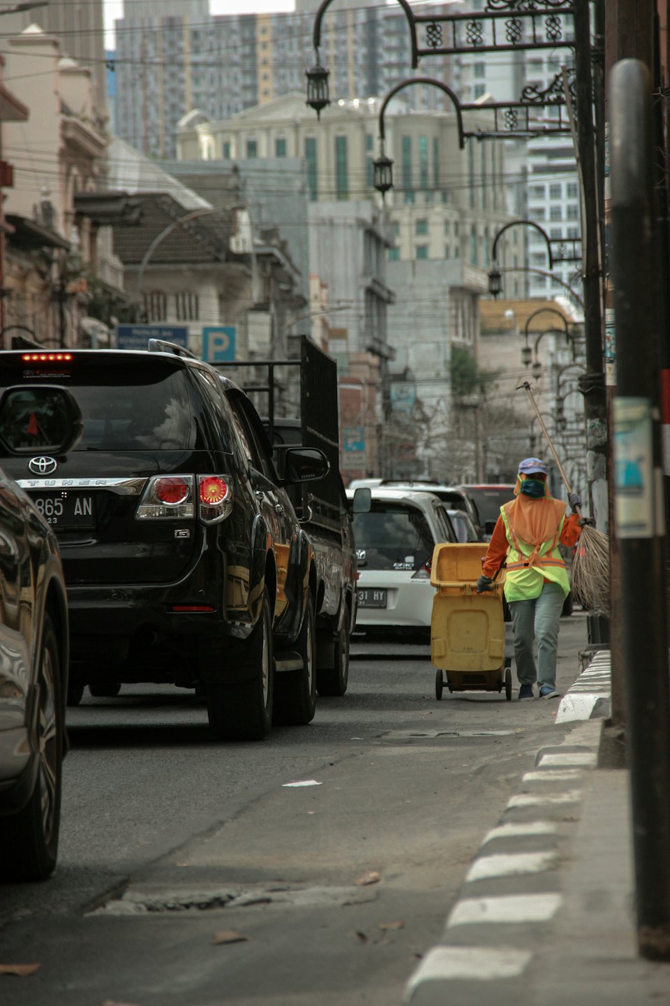 Worker pushes trash bin and carries broom on sidewalk.